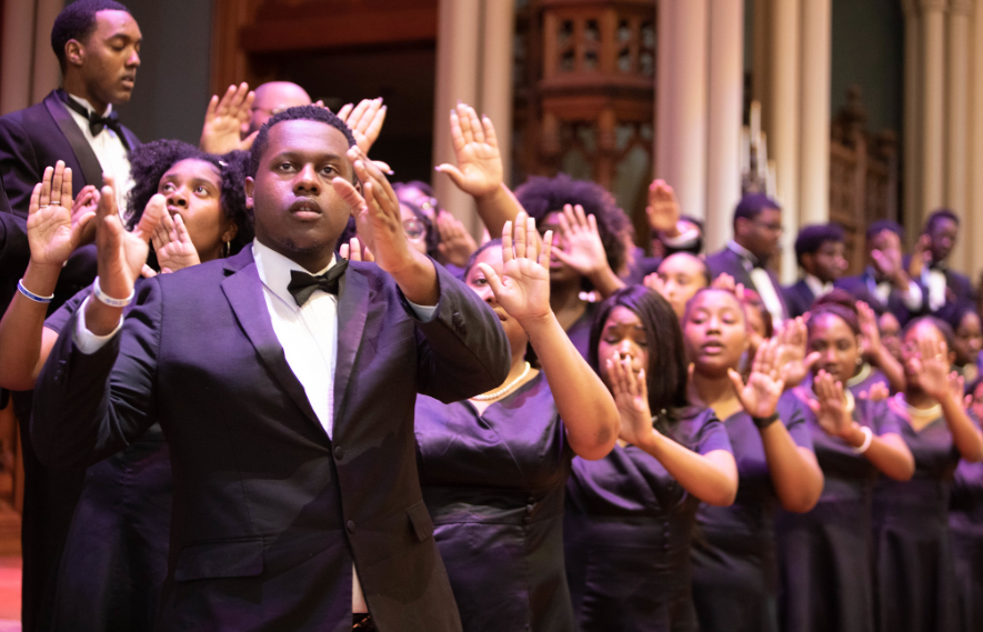 Several choir members in black choral wear sing with their hands raised in the air. 