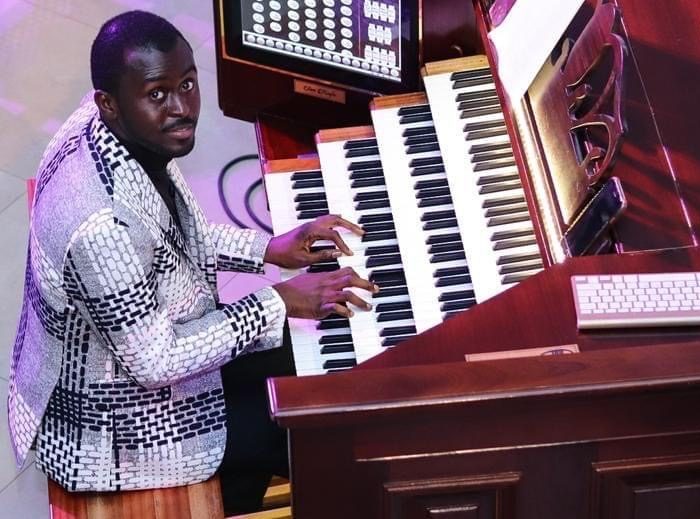 Jospeh sits at a wooden organ wearing a black and gray jacket. 